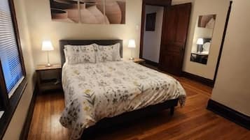 A bedroom with a wooden floor, a bed with a floral bedspread, two bedside lamps, a ceiling fan, and a wall-mounted artwork.