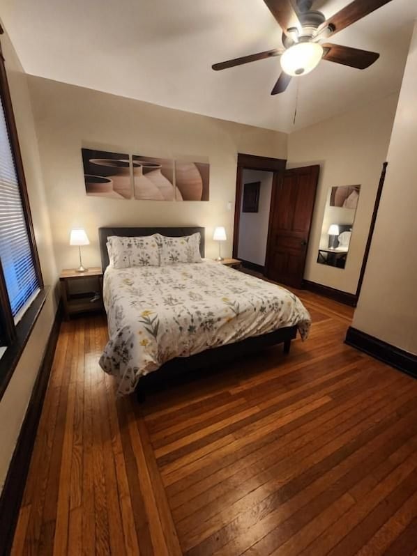 A bedroom with a wooden floor, a bed with a floral bedspread, two bedside lamps, a ceiling fan, and a wall-mounted artwork.