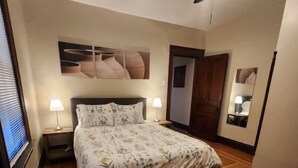 A bedroom with a wooden floor, a bed with floral bedding, two bedside lamps, a ceiling fan, and artwork on the walls. - Cleneay #1, Private 3 Bed near Xavier & Downton (Cincinnati)