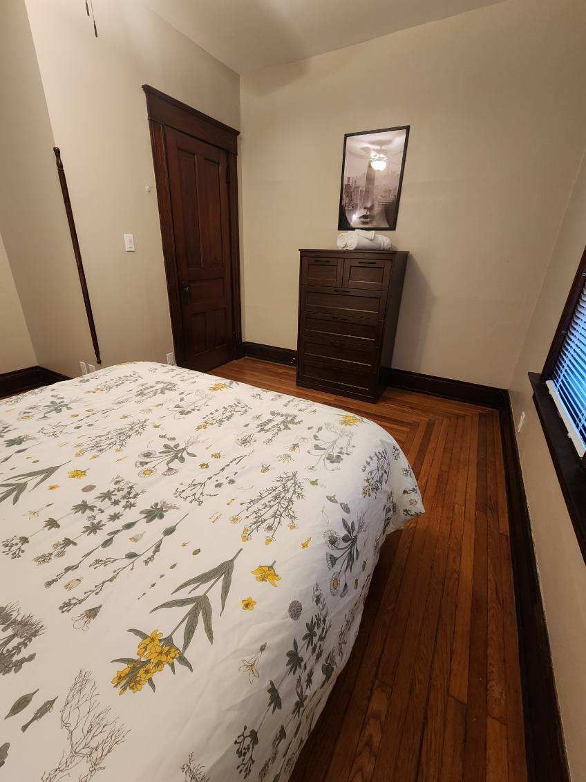 A bedroom with a floral bedspread, a wooden dresser, a framed picture on the wall, and a window with blinds.