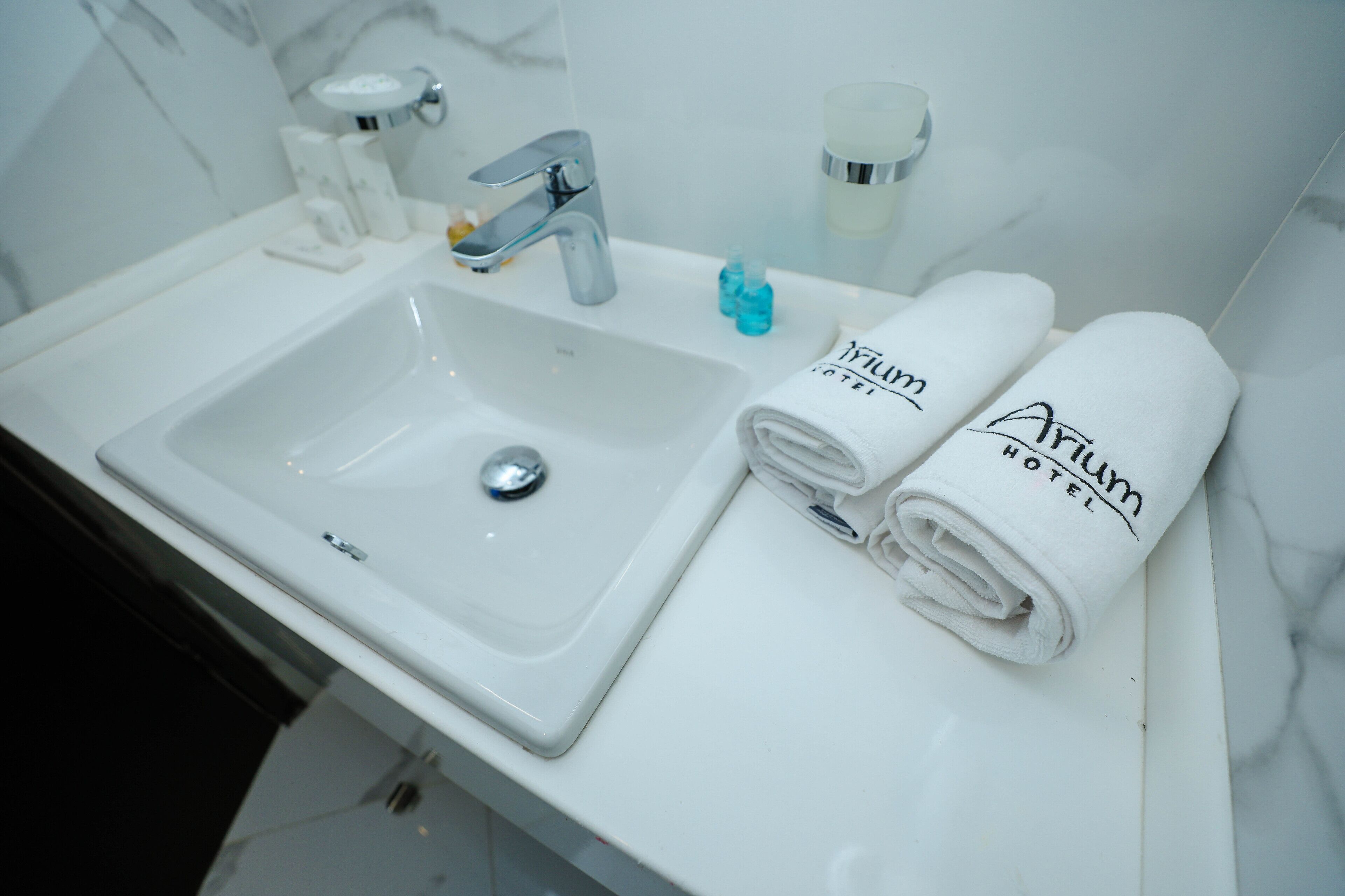 A white bathroom sink with a chrome faucet, two rolled towels with "Arion Hotel" branding, and a small blue bottle of soap.