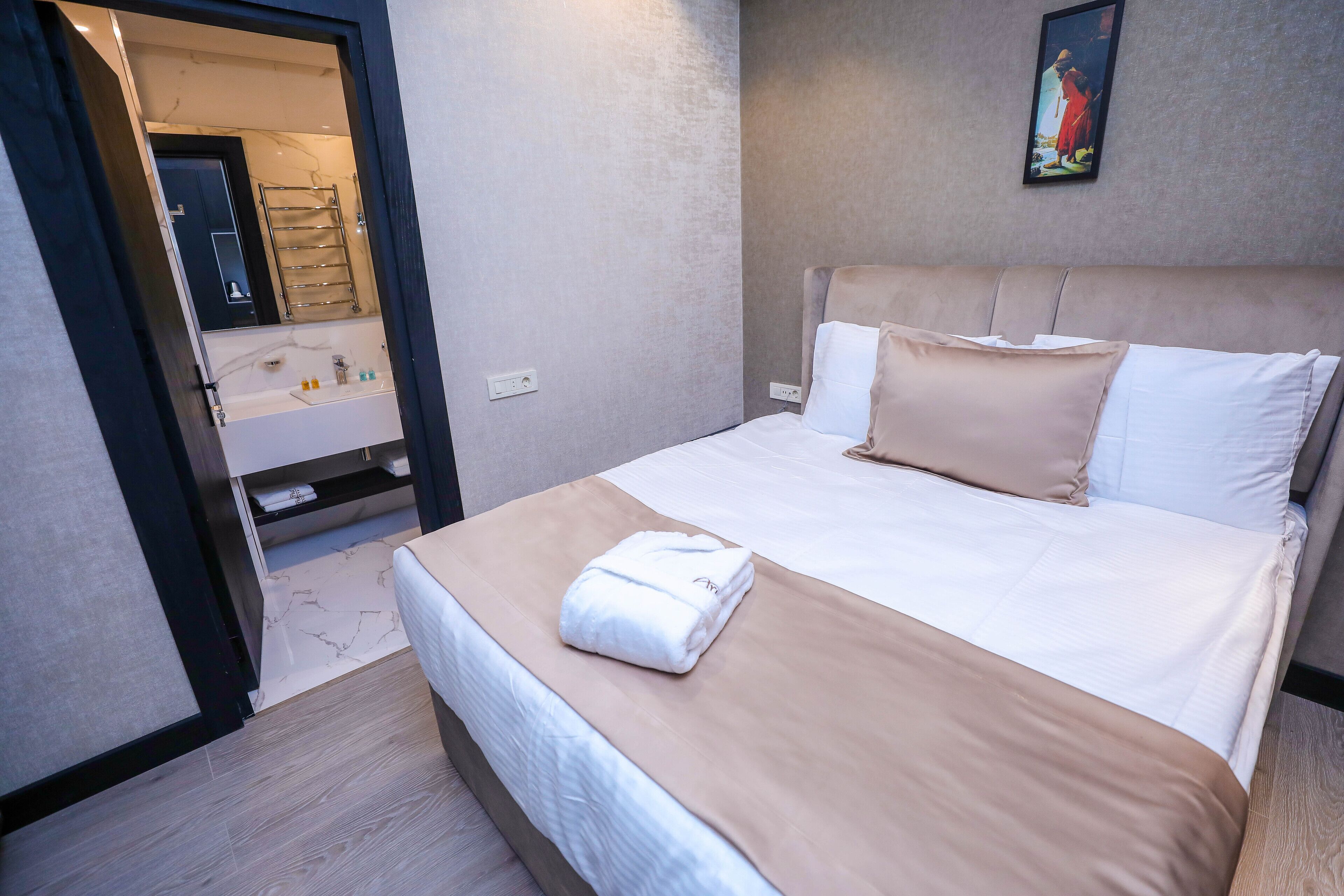 Una cama bien tendida con edredón color crema y blanco, una toalla blanca sobre la cama y un cuadro colgado en la pared.