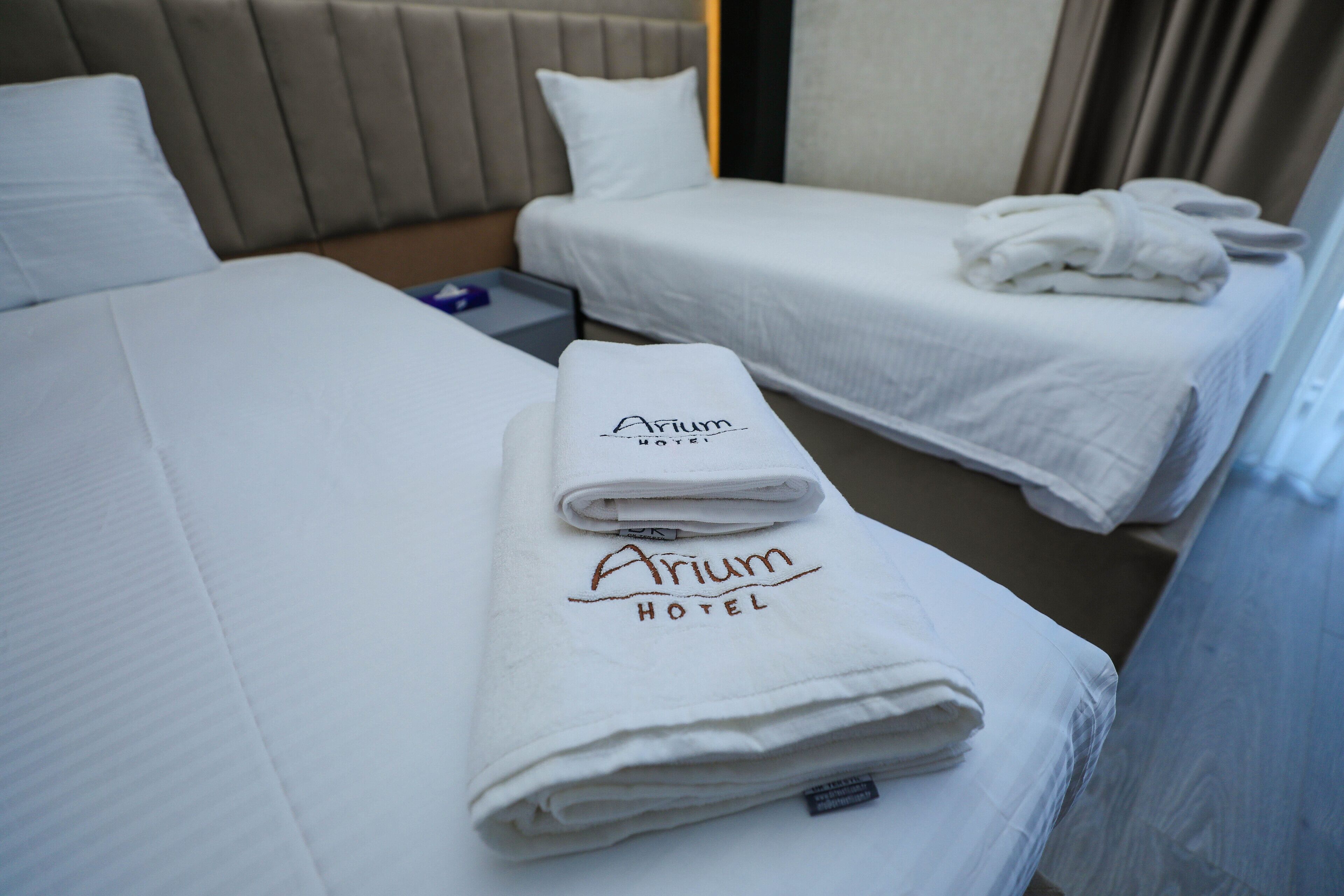 A hotel room with two beds, each with a folded white towel and a pillow. The room has a neutral color scheme with beige and white tones.