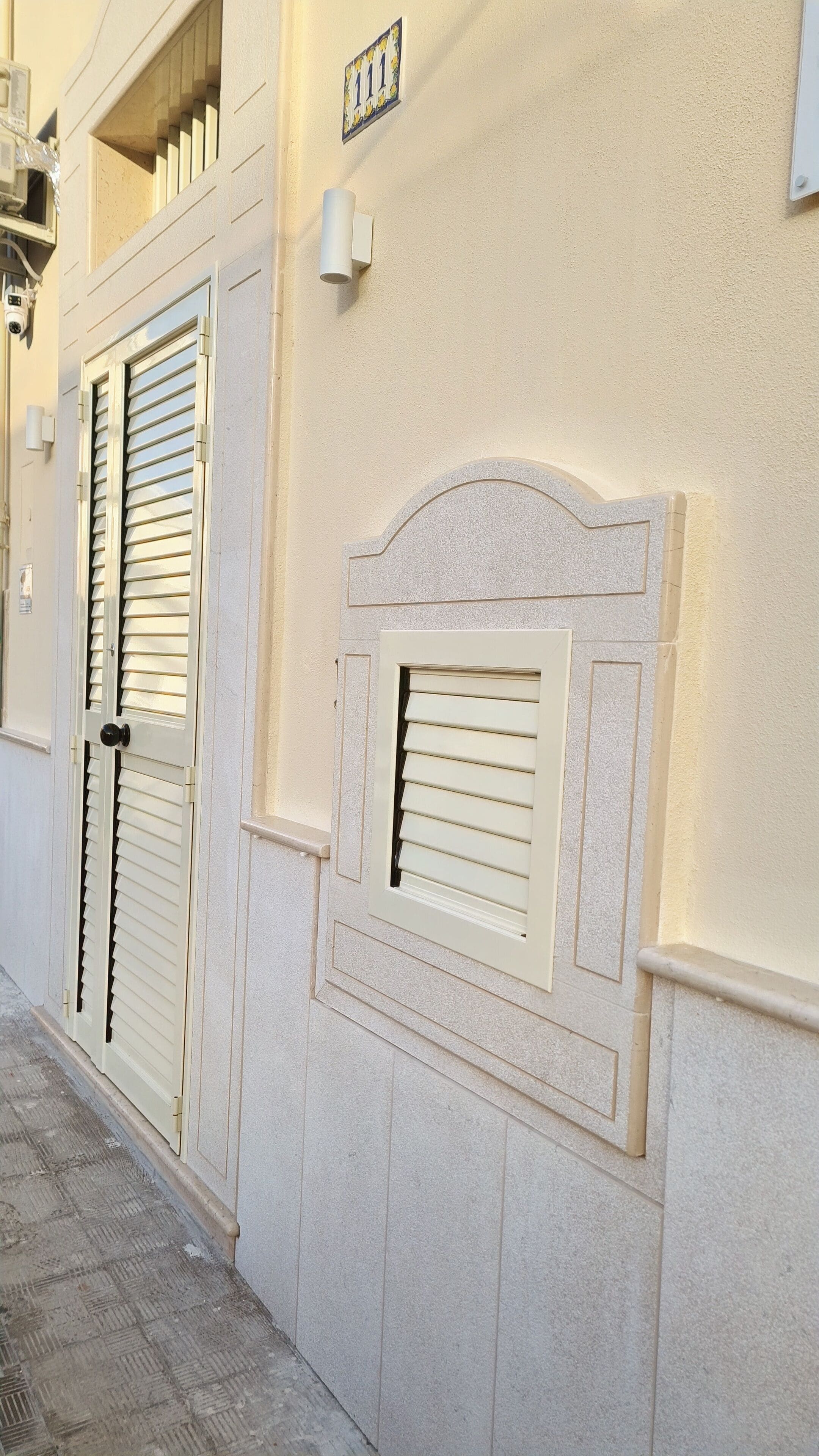 Eine Gebäudefassade in Beige, eine Tür mit schwarzem Griff und ein Fenster mit Rollläden.