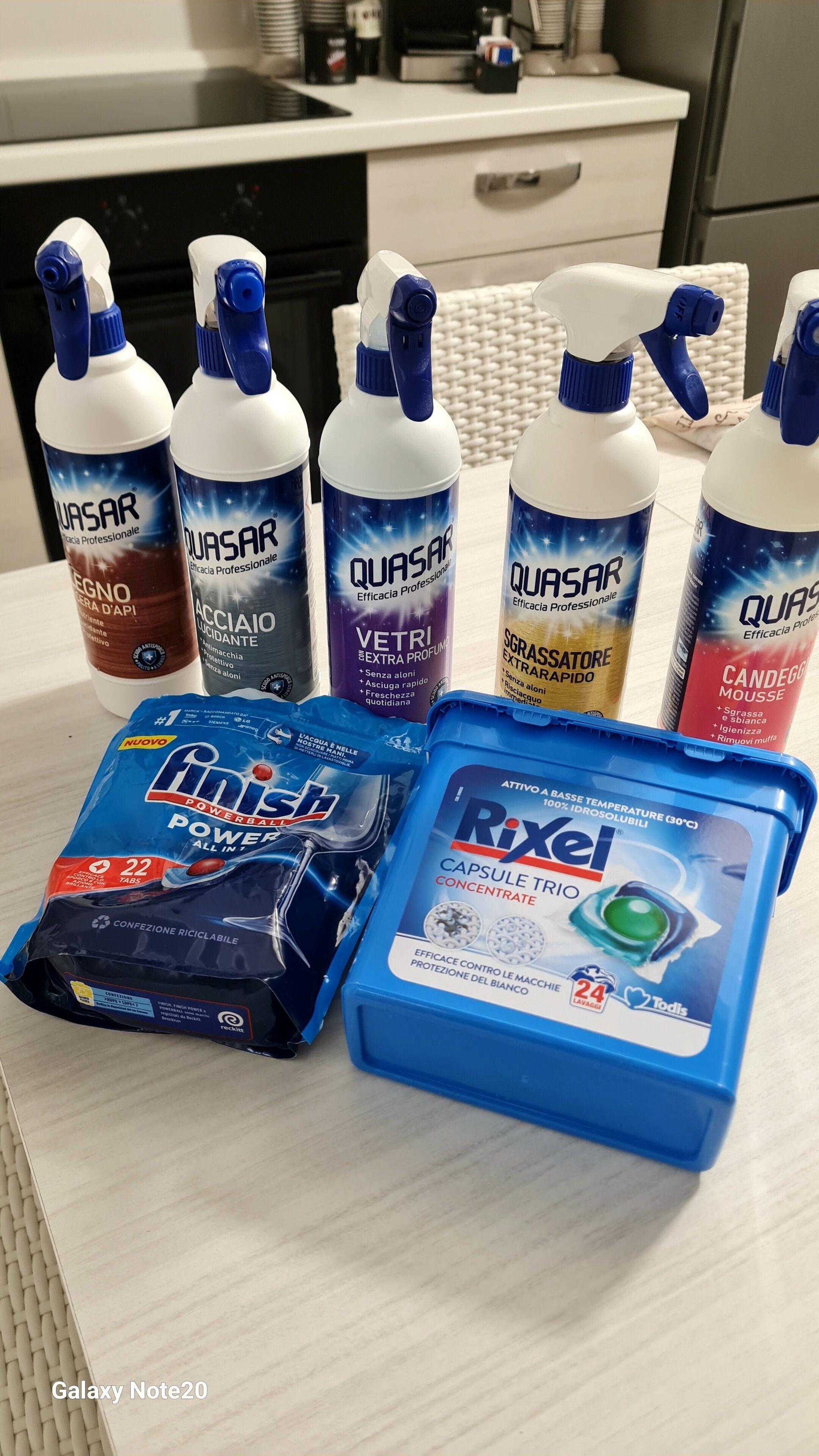 Un plan de travail de cuisine avec divers produits de nettoyage, notamment des bouteilles de la marque Quasar et un récipient bleu contenant du produit concentré Rixe.
