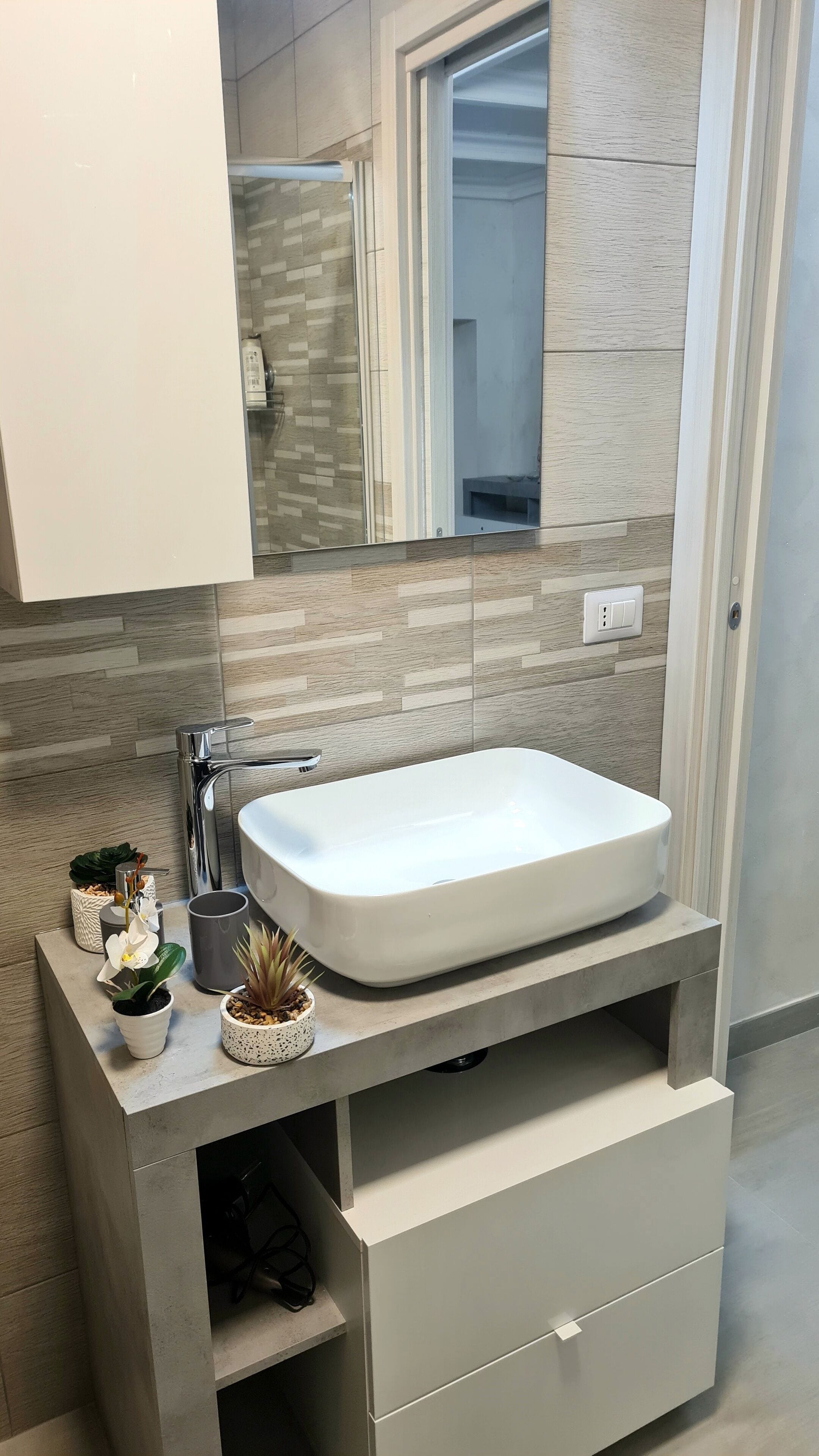 A modern bathroom with a rectangular white sink, a mirror above it, and a tiled backsplash. There are two small potted plants on the countertop.