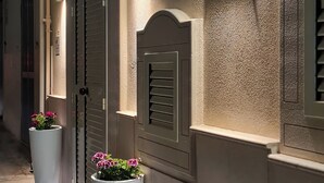 Una calle estrecha con un edificio de fachada texturizada, una puerta con un panel decorativo y dos macetas grandes con flores.