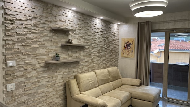 Un salon moderne avec un mur à l’aspect pierre, un canapé d’angle beige, une table à manger et une vue sur un toit recouvert de tuiles rouges, visible à travers une porte-fenêtre coulissante.