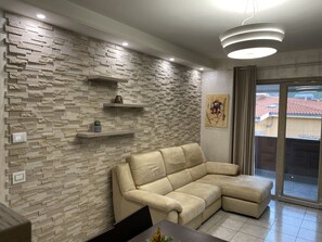 A modern living room with a stone-textured wall, a beige sectional sofa, a dining table, and a view of a red-tiled roof through a sliding glass door. - Apartment 4 minutes walk from the beach. (Bosa)