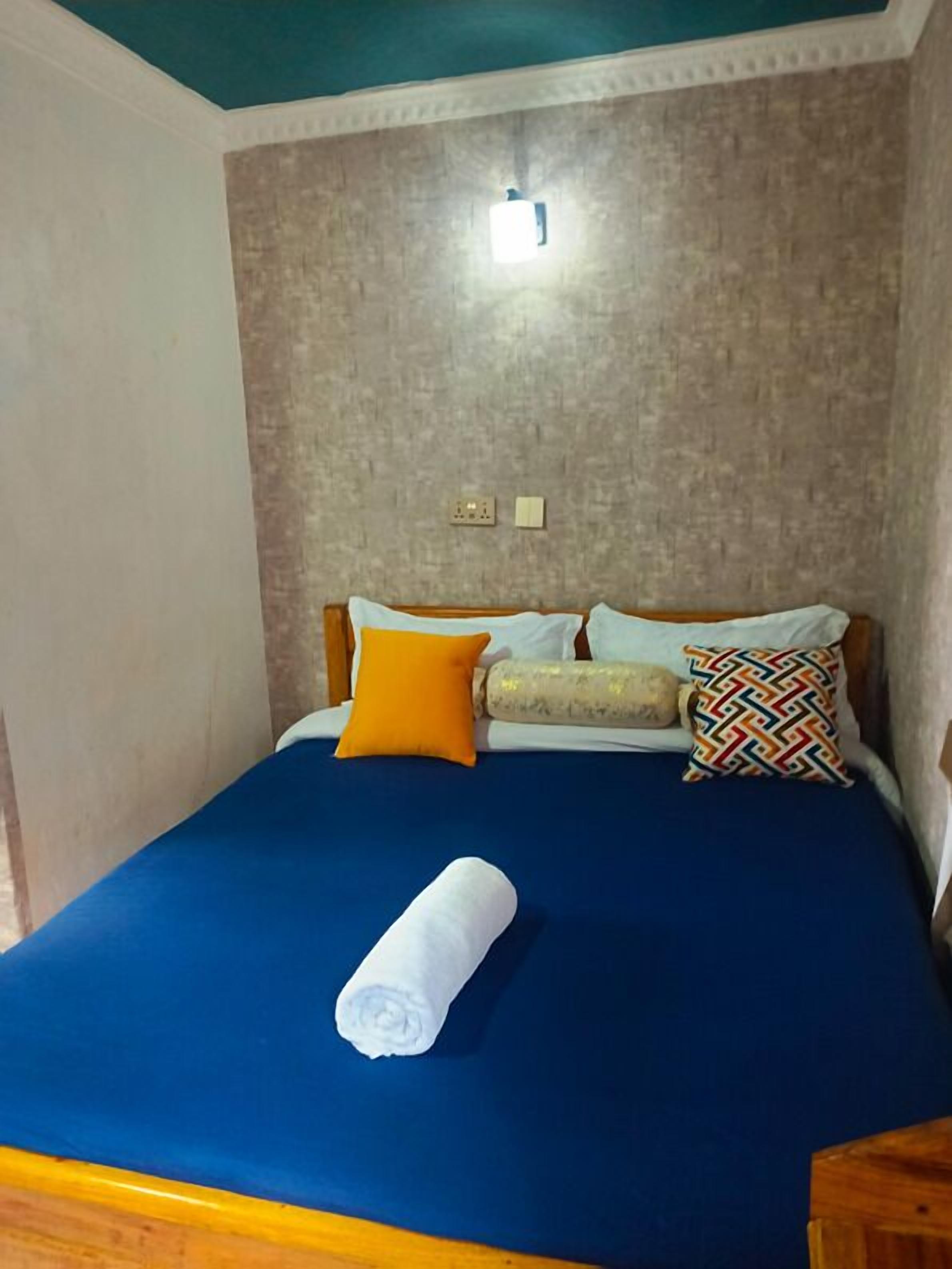 A bed with a blue mattress, two pillows, a rolled white towel, and a patterned pillow.
