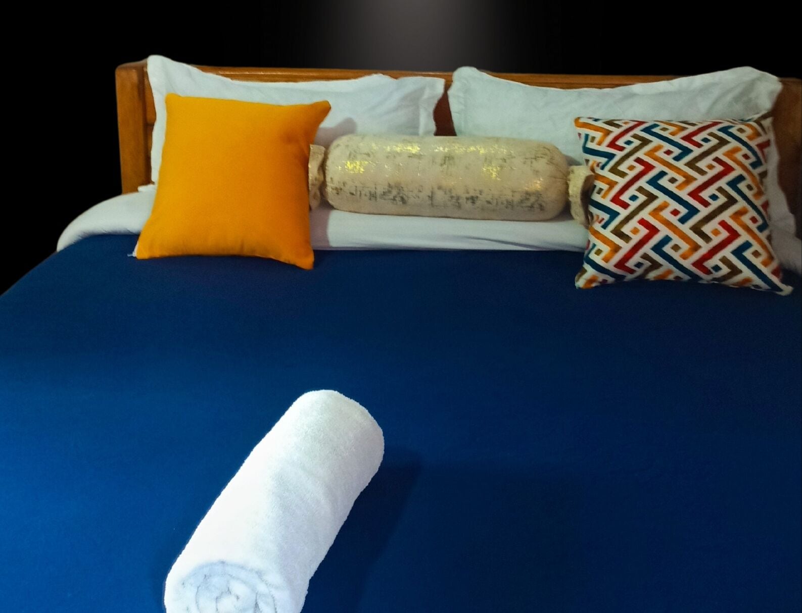 A bed with a blue bedspread, a white towel, and three pillows in different colors and patterns.