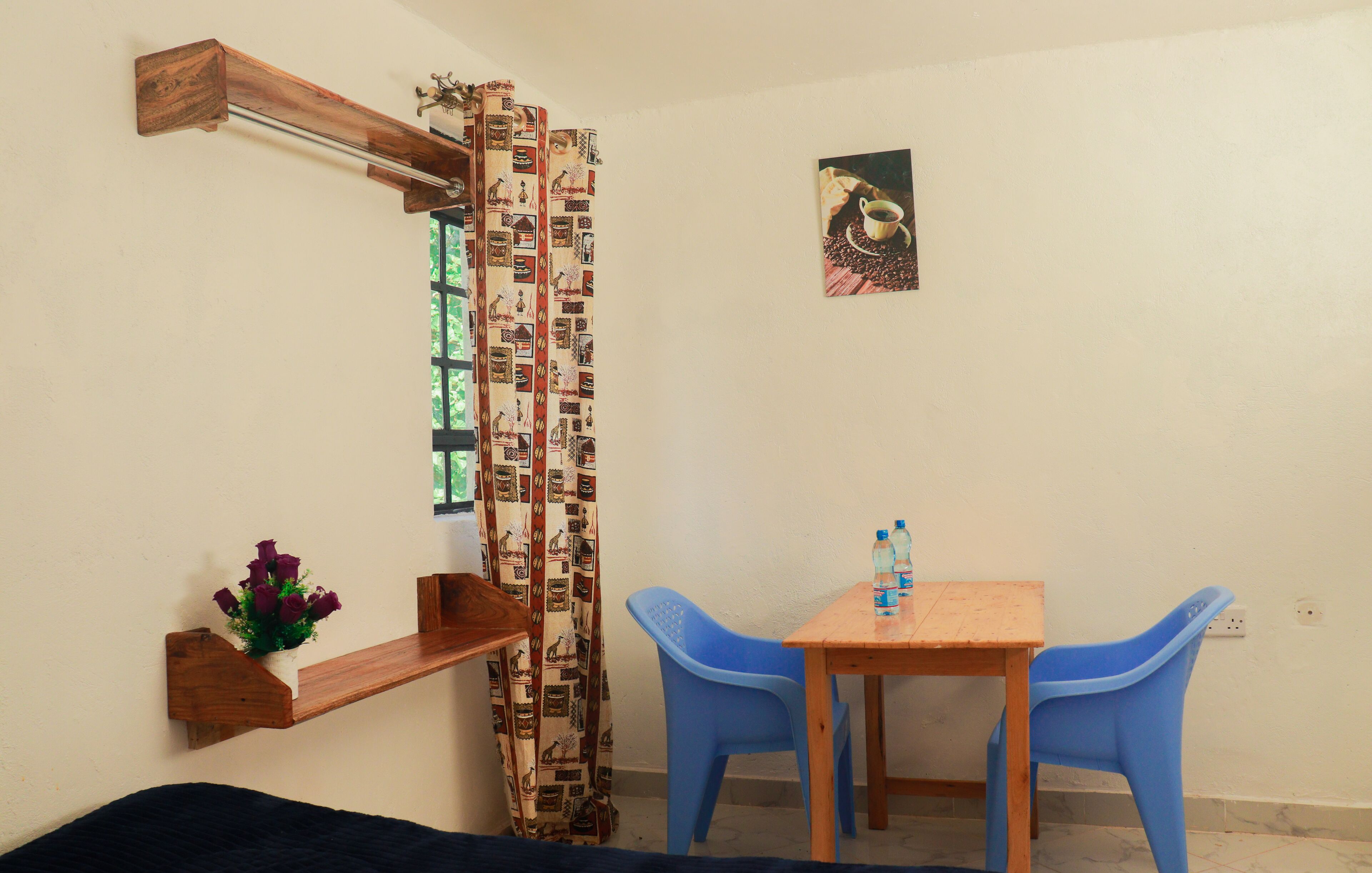 A small room with a wooden shelf, a blue plastic chair, a wooden table, a curtain with a pattern, and a picture on the wall.