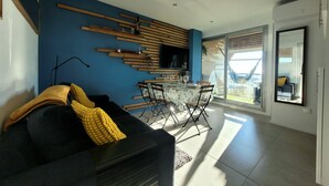 Ein modernes Wohnzimmer mit einer schwarzen Couch, einem Esstisch mit Stühlen, einem an einer blauen Wand montierten Fernseher und einem großen Fenster mit Blick ins Freie.