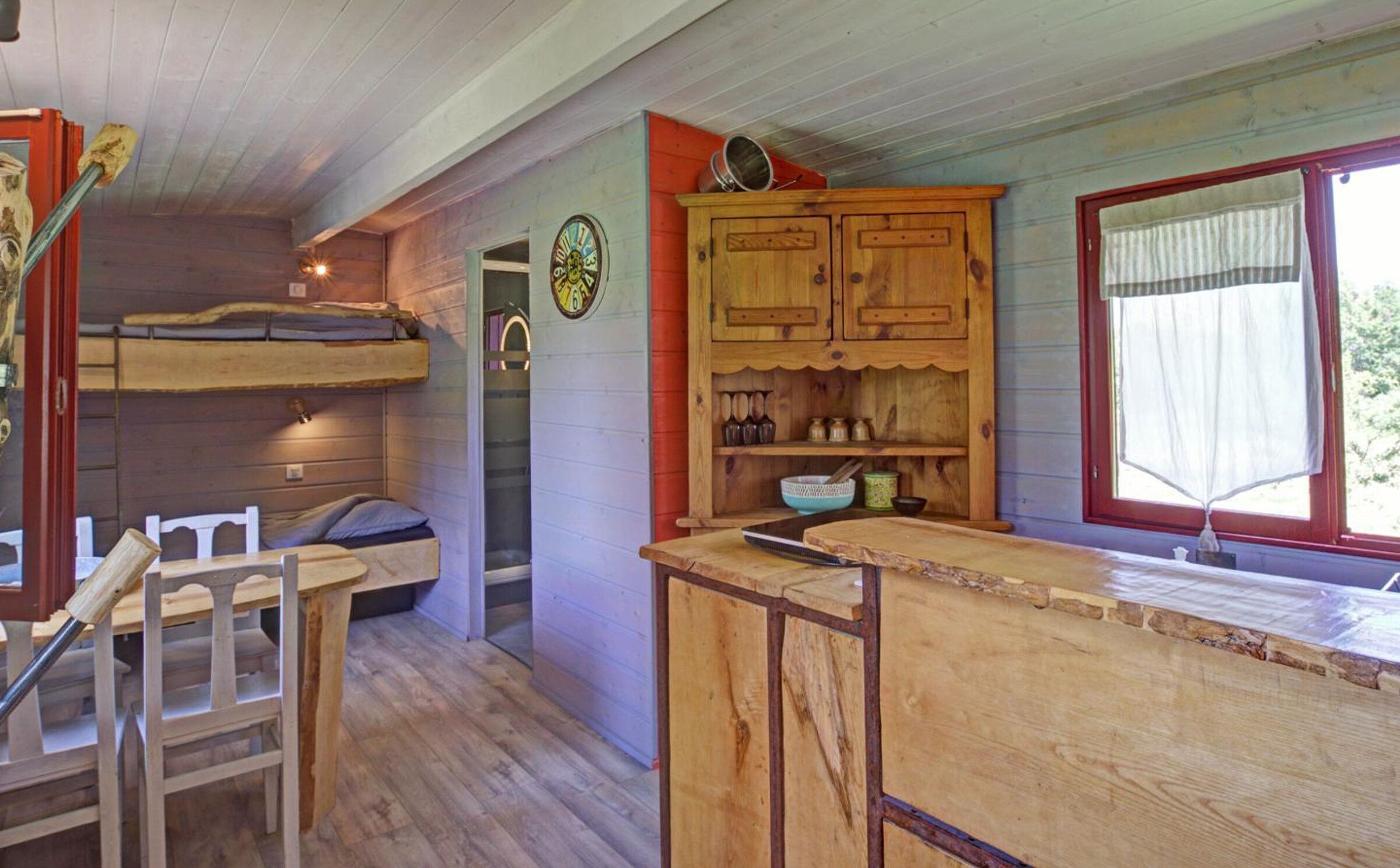 A rustic cabin interior with a wooden dining table, chairs, a kitchen area with wooden cabinets, and a window with curtains.