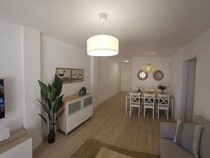 A modern living room with a dining area, featuring a sofa, a dining table with chairs, a hanging light fixture, and a potted plant.