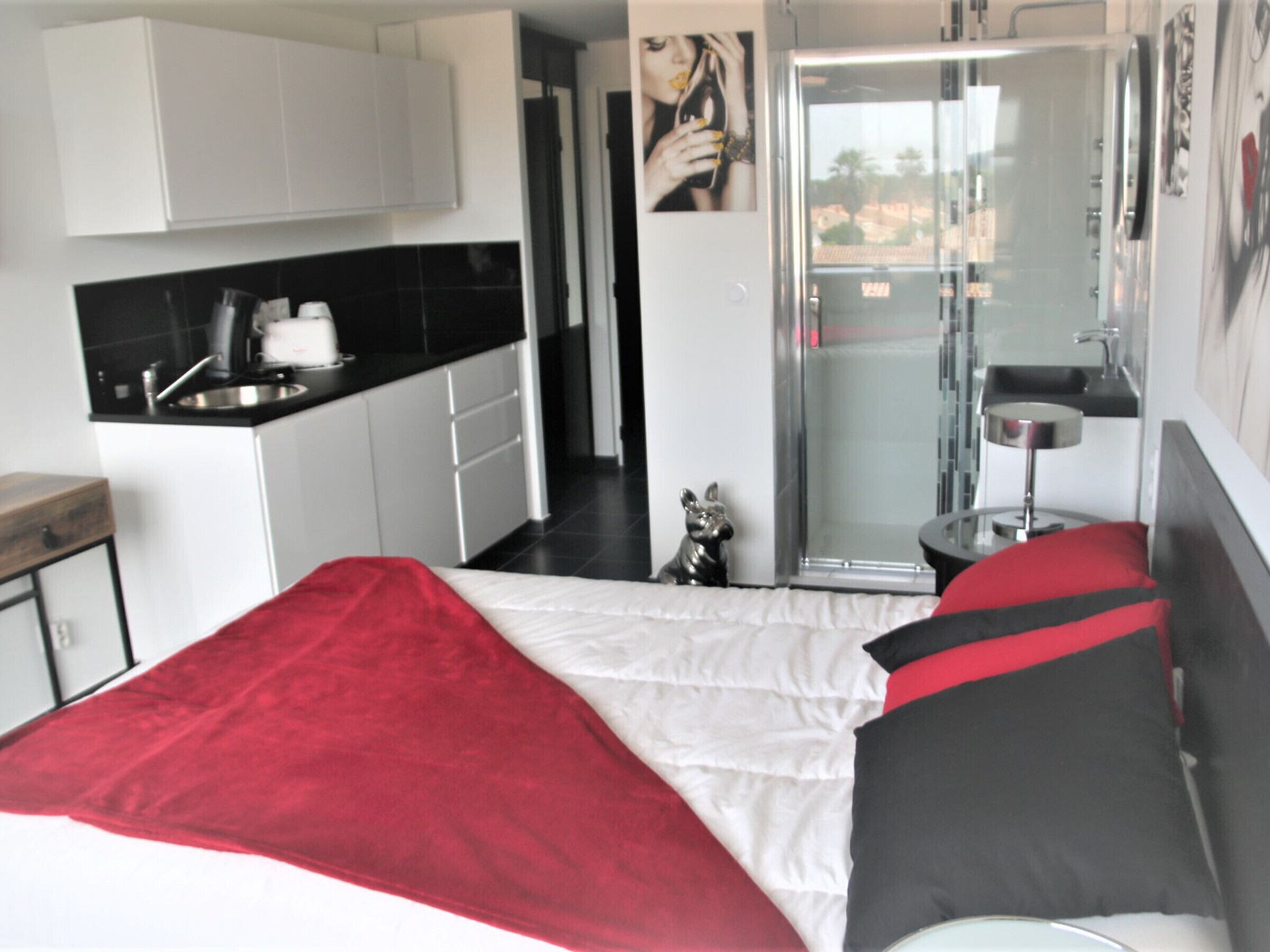 A bedroom with a bed, a red and white blanket, a small dog figurine, a bathroom with a glass shower, and a kitchen area.