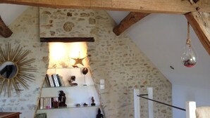 A rustic room with exposed wooden beams, a stone wall, and a shelf with books and decorative items. - Charming Renovated Cottage with Pool and Art near Le Mans (Lucé-sous-Ballon)