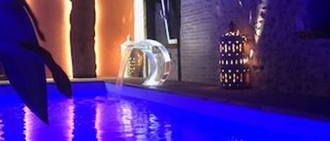 Un área de piscina interior moderna con estructura de vidrio transparente, fuente de agua e iluminación decorativa.