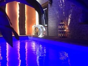 A modern indoor pool area with a clear glass structure, a water feature, and decorative lighting. - Charming Renovated Cottage with Pool and Art near Le Mans (Lucé-sous-Ballon)