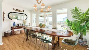 A dining area with a wooden table, four chairs, a chandelier, a large mirror, and a potted plant. - Pink Pearl - Where Mr Ocean Blue Meets Ms Posh Pink! (Surfside Beach)