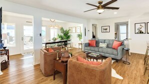 A spacious living room with a gray sofa, brown armchairs, and a wooden coffee table. A ceiling fan and a dining area with a glass table are visible in the background. - Pink Pearl - Where Mr Ocean Blue Meets Ms Posh Pink! (Surfside Beach)