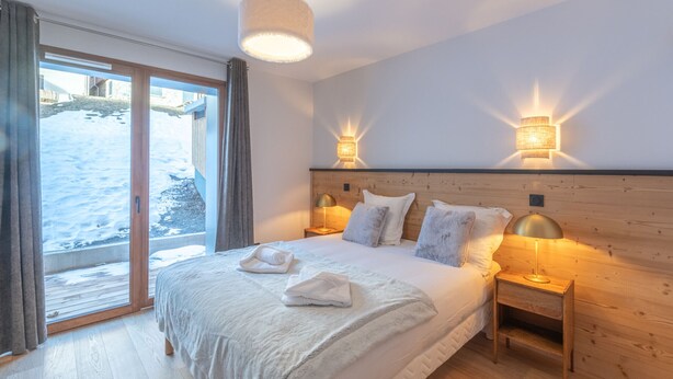 A bedroom with a large bed, wooden headboard, two bedside lamps, and a view of the outside. - New apartment 108m², center Huez, parking, wifi, 6 guests (Huez)