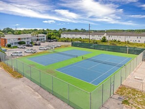 Un complexe de tennis comprenant plusieurs courts, entouré d’une clôture et situé à côté d’un parking où sont stationnées plusieurs voitures.