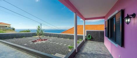 A covered outdoor area with a tiled floor, a garden with small plants, and a view of a building with orange tiles.