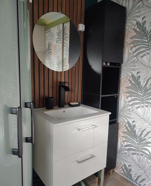 Un baño con tocador blanco, espejo redondo, mueble negro y papel tapiz con estampado de hojas.