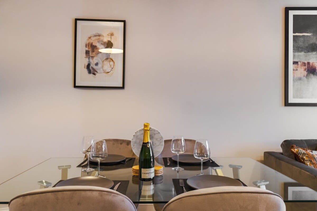 Een eettafel met een champagnefles, twee glazen en een lijst met een abstract schilderij aan de muur.