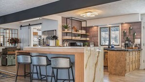 A modern kitchen with a marble island, bar stools, wooden cabinets, and a large window. - Spacious 4BR, Games, Pool, Theater & Private Yard! (Omaha)