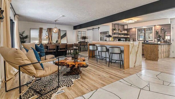 A modern living room with a sofa, a coffee table, and a dining area. There is a kitchen with a bar and a large window. - Spacious 4BR, Games, Pool, Theater & Private Yard! (Omaha)