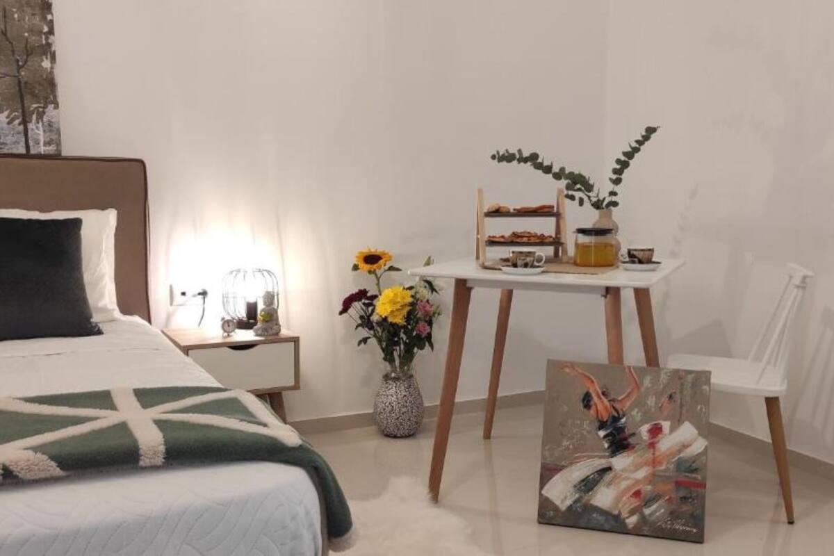 Een slaapkamer met een bed, nachtkastje, lamp, vaas met bloemen en een schilderij.
