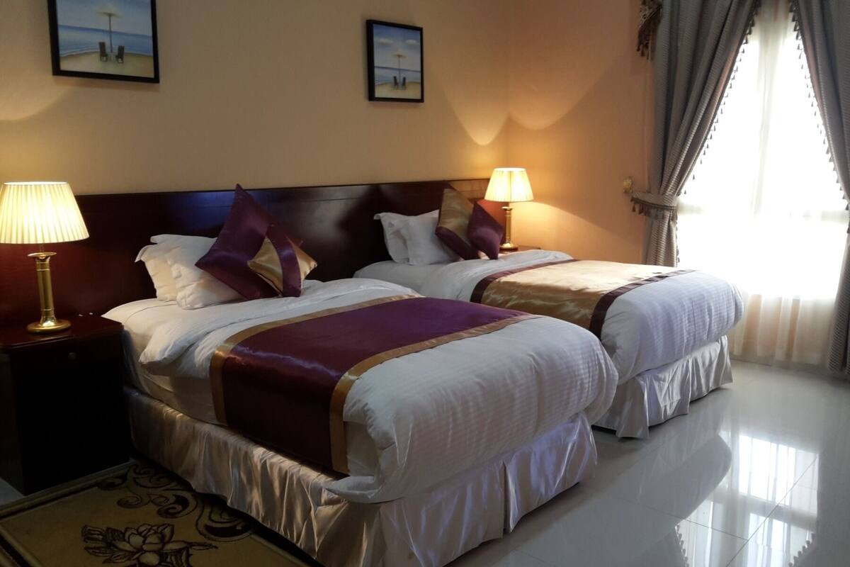 A hotel room with two beds, each with a headboard, bedside lamps, and a view of the beach through the window.
