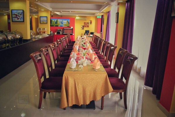 A long dining table with chairs, set for a meal, in a room with yellow walls and a red accent wall. - The Royal Garden Hotel (Sohar)