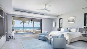 A modern bedroom with a large window overlooking a beach, a bed, a desk, and a chair.