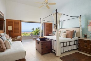 A bedroom with a four-poster bed, a wooden chest of drawers, and a view of the ocean.
