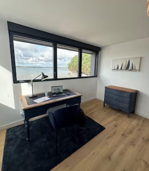 A room with a desk, chair, and a large window with a view of the sea.