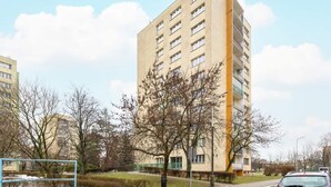 Un edificio alto de apartamentos color beige, con varios balcones, rodeado de árboles sin hojas y un cielo despejado.