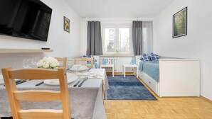 Una habitación compacta con cama, una mesita con silla, televisor montado en la pared y ventana con cortinas.