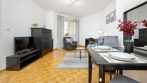 A modern living room with a flat-screen TV, dark furniture, a dining table set for two, and a large window with curtains. - Functional Apartment | Parking, Workspace (Warszawa)