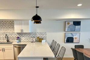 Una cocina moderna con encimera de mármol, una lámpara colgante negra, salpicadero con estampado en zigzag y una zona de comedor con sillas de metal.