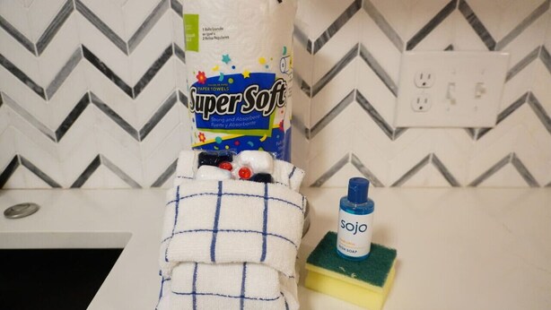 A bathroom with a white sink, a towel, a box of SuperSoft paper towels, a bottle of dish soap, and a green sponge.