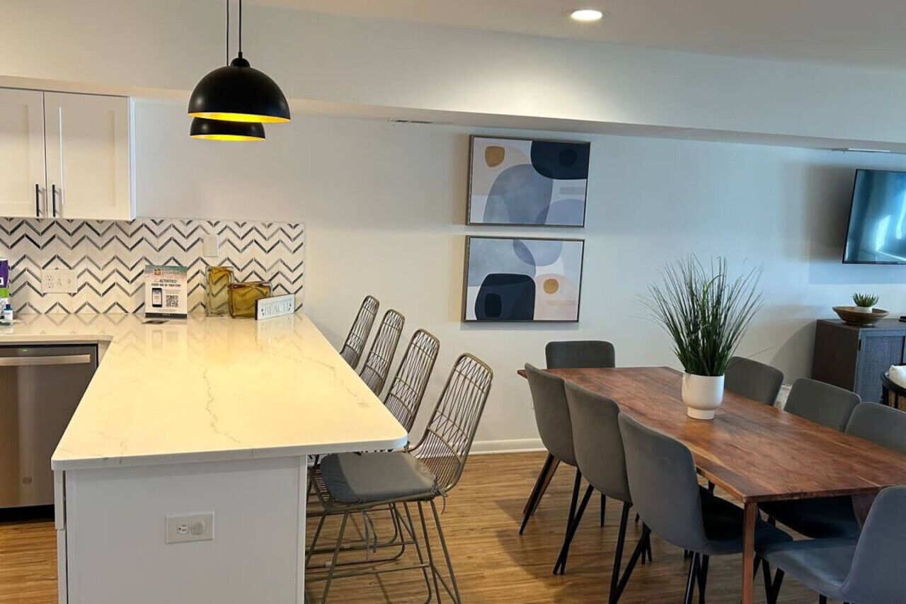 Una cocina moderna con encimera de mármol blanco, lavavajillas integrado y una lámpara colgante negra. En la pared, hay dos cuadros abstractos enmarcados, una mesa de comedor con sillas y una planta en maceta sobre la mesa.