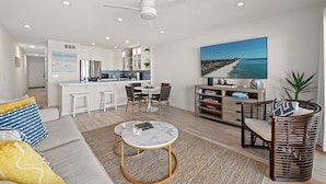 Un salon moderne avec un canapé, une table basse et un coin repas avec des chaises. On y trouve une cuisine avec des meubles blancs et une grande fenêtre. Un tableau représentant une plage est accroché au mur.