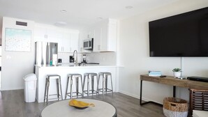 A modern kitchen with white cabinets, a stainless steel refrigerator, and a flat-screen TV mounted on the wall. There are four bar stools and a round table with a yellow bowl in the center. - Seamist 7 ~ Beachfront on 30A (Seacrest)