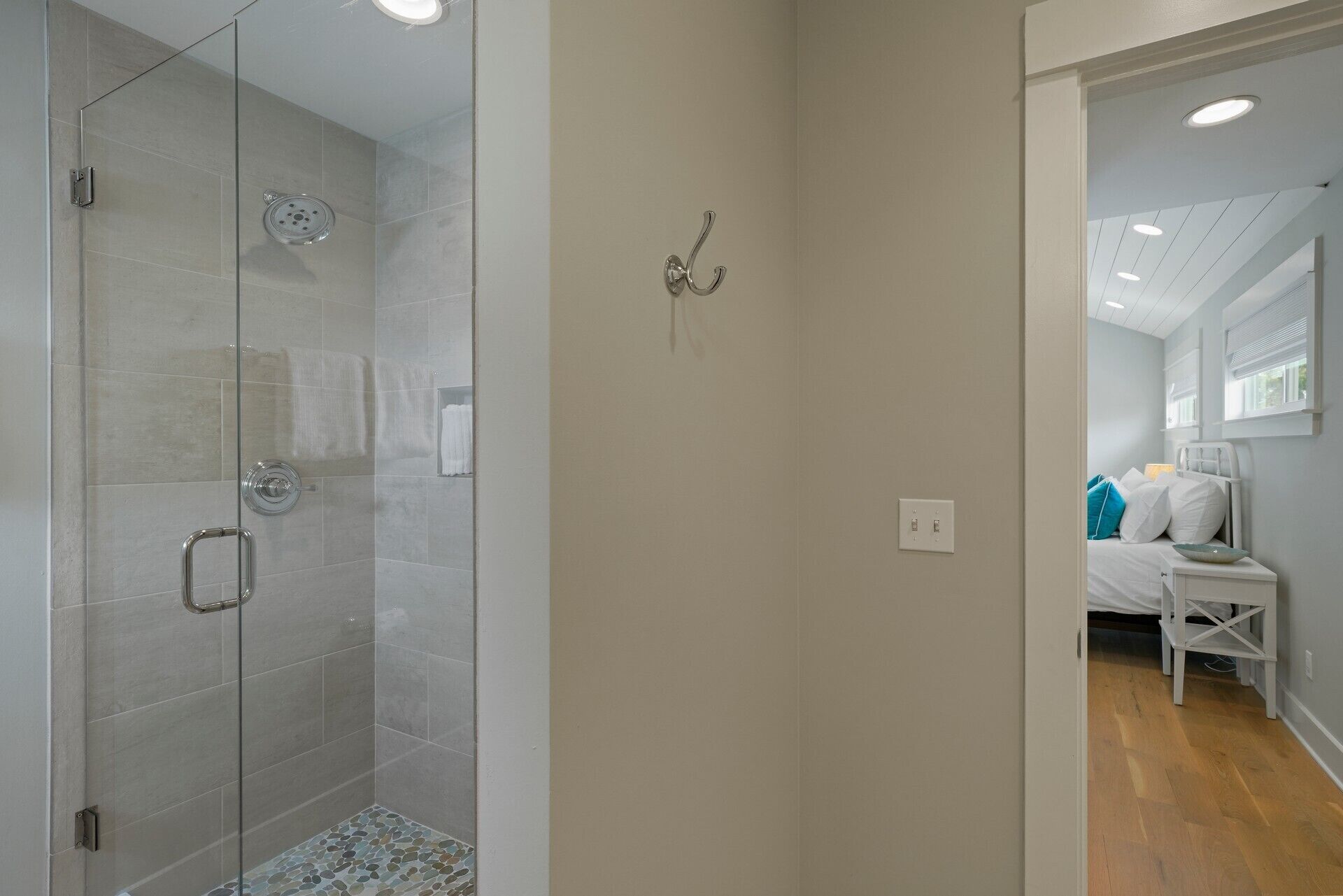 A modern bathroom with a glass-enclosed shower, a towel rack, and a bed visible through an open door.