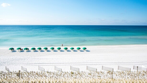 Une plage de sable blanc bordée d’une mer d’un bleu limpide. Des parasols verts sont alignés sur le sable.