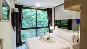 A hotel room with a bed, a TV, a window with curtains, and a view of greenery outside.