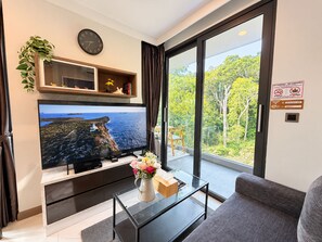 A modern living room with a large flat-screen TV, a glass coffee table, a sofa, and a view of a lush green landscape through large sliding glass doors.